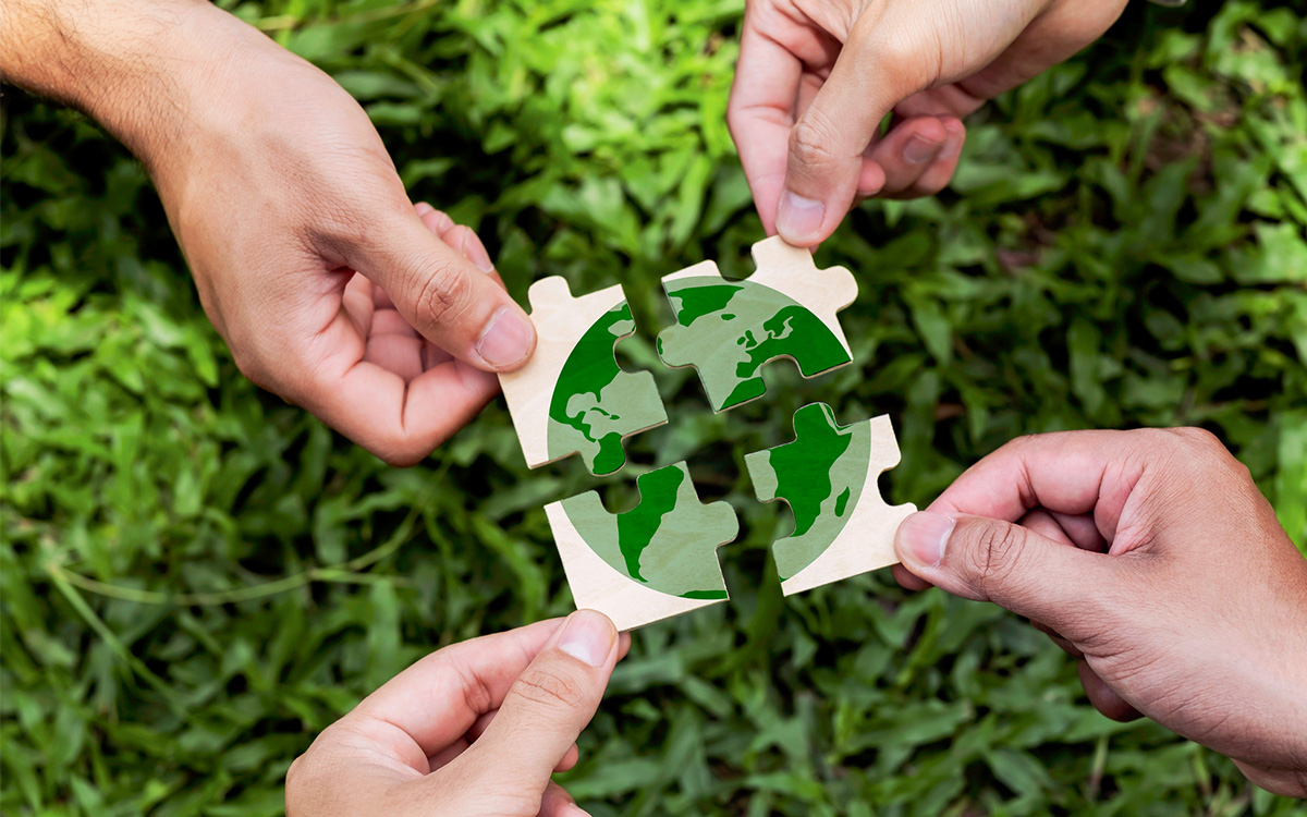 Four hands hold puzzle pieces that form an image of the Earth over green grass.