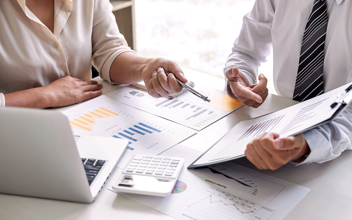 Two people in business attire review documents with charts and graphs at a desk, using a laptop, calculator, and clipboard.