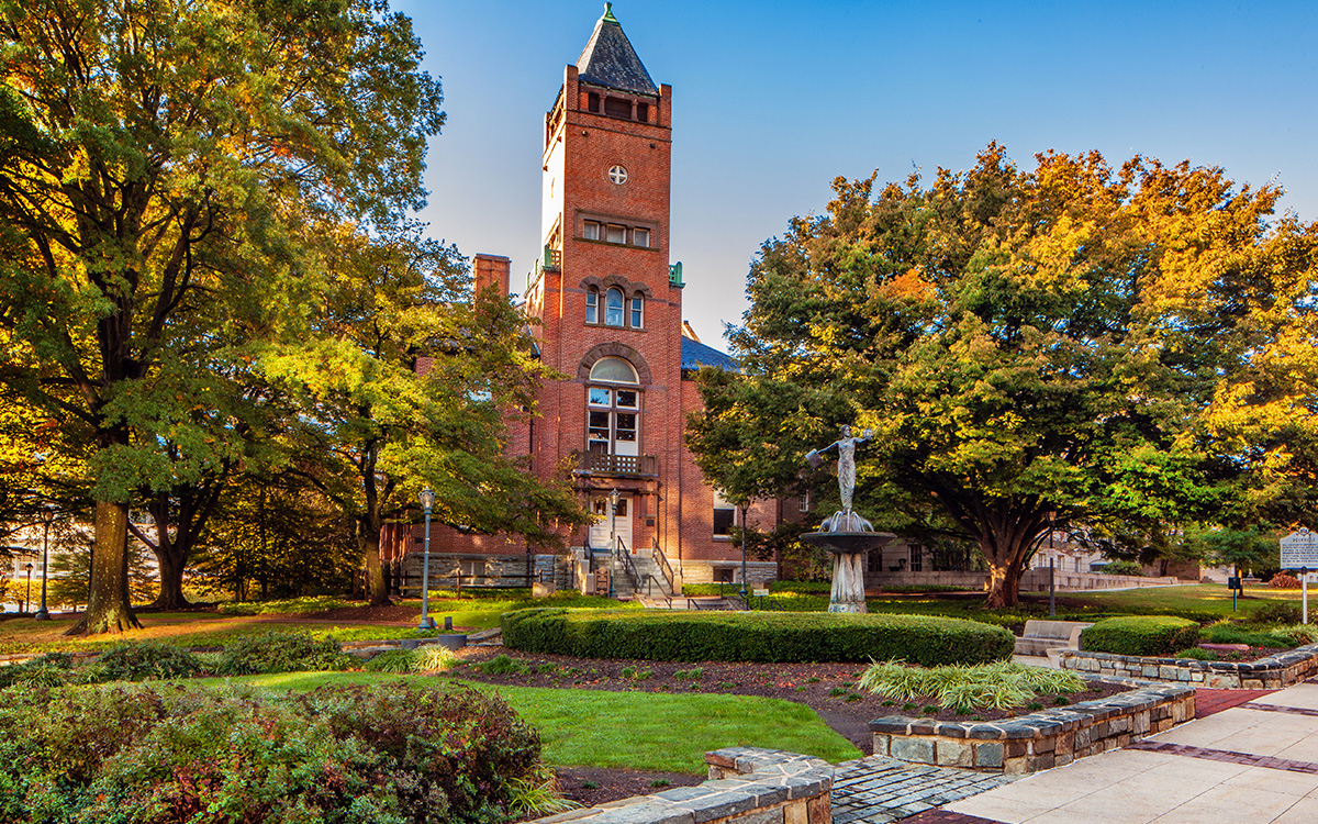 A historic red brick building with a tall clock tower, surrounded by trees and landscaped gardens under a clear blue sky.