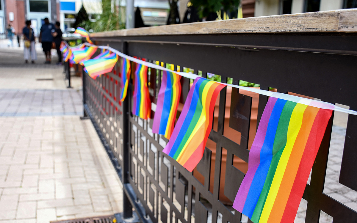 Small rainbow pride flags are strung along a black metal fence on a city sidewalk. Small rainbow pride flags are strung along a black metal fence on a city sidewalk.