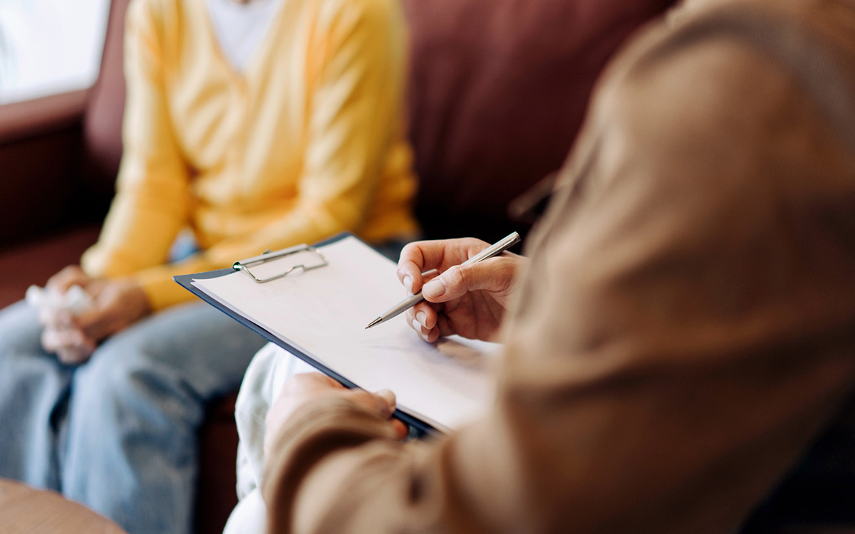A person holding a clipboard and pen sits across from another person in a yellow sweater, suggesting a counseling or interview session. A person holding a clipboard and pen sits across from another person in a yellow sweater, suggesting a counseling or interview session.