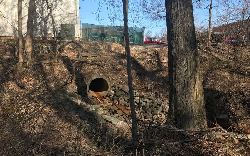 A large metal drainage pipe emerges from a hillside surrounded by rocks and trees, with a building and fencing visible in the background.