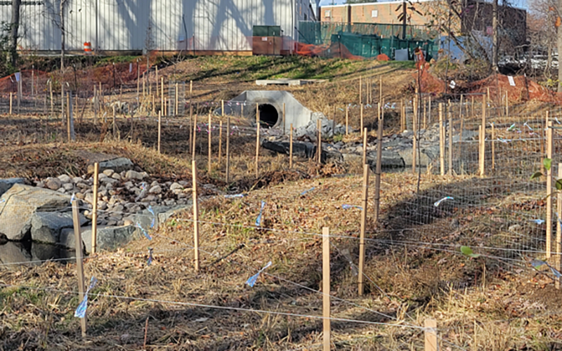 A field with dry grass, wooden stakes, mesh fencing, rocks, and a concrete drainage pipe in the background near industrial buildings.