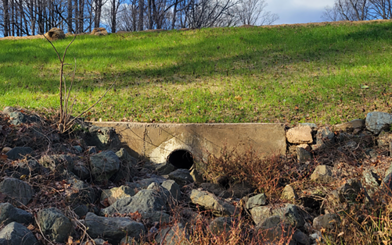 A concrete drainage pipe emerges from a rocky slope below a grassy embankment with leafless trees in the background.