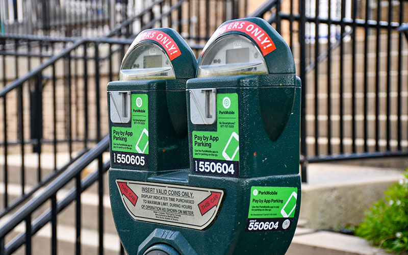 A close-up of two green parking meters with stickers indicating pay-by-app options, situated in front of a metal railing and stairs.