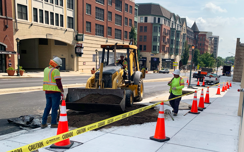 Construction workers use a backhoe to move soil on a city street. Traffic cones and caution tape mark the area for safety. Nearby buildings and parked cars are visible.