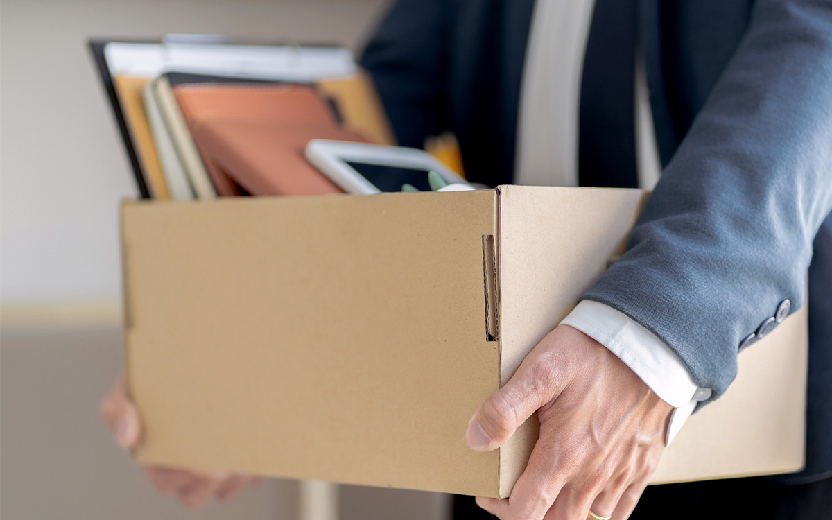 A person wearing a suit holds a cardboard box filled with office supplies, documents, and personal items.