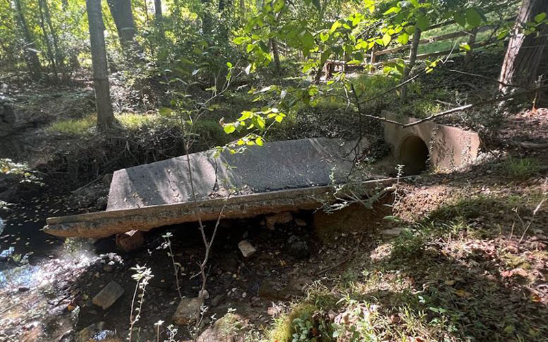 A broken concrete bridge slab lies partially collapsed over a small creek in a wooded area, with a nearby drainage pipe visible.