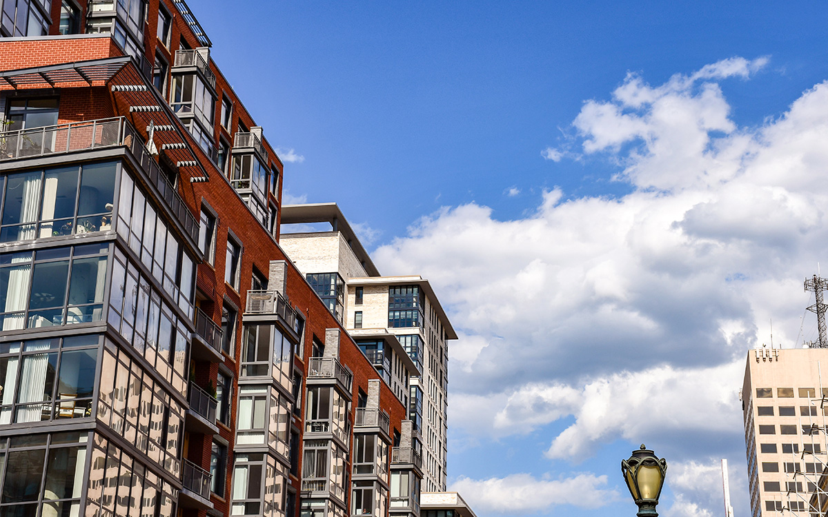 Modern apartment buildings with large windows are shown against a bright blue sky with scattered clouds. A streetlamp and a tall office building are visible in the foreground and background. Modern apartment buildings with large windows are shown against a bright blue sky with scattered clouds. A streetlamp and a tall office building are visible in the foreground and background.