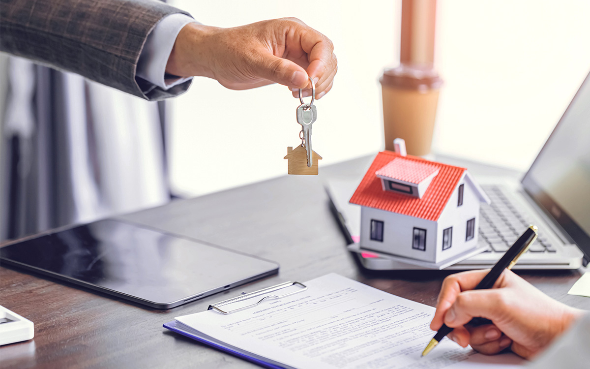 A person hands over keys with a house-shaped keychain while another person signs documents at a desk with a model house, laptop, and tablet. A person hands over keys with a house-shaped keychain while another person signs documents at a desk with a model house, laptop, and tablet.