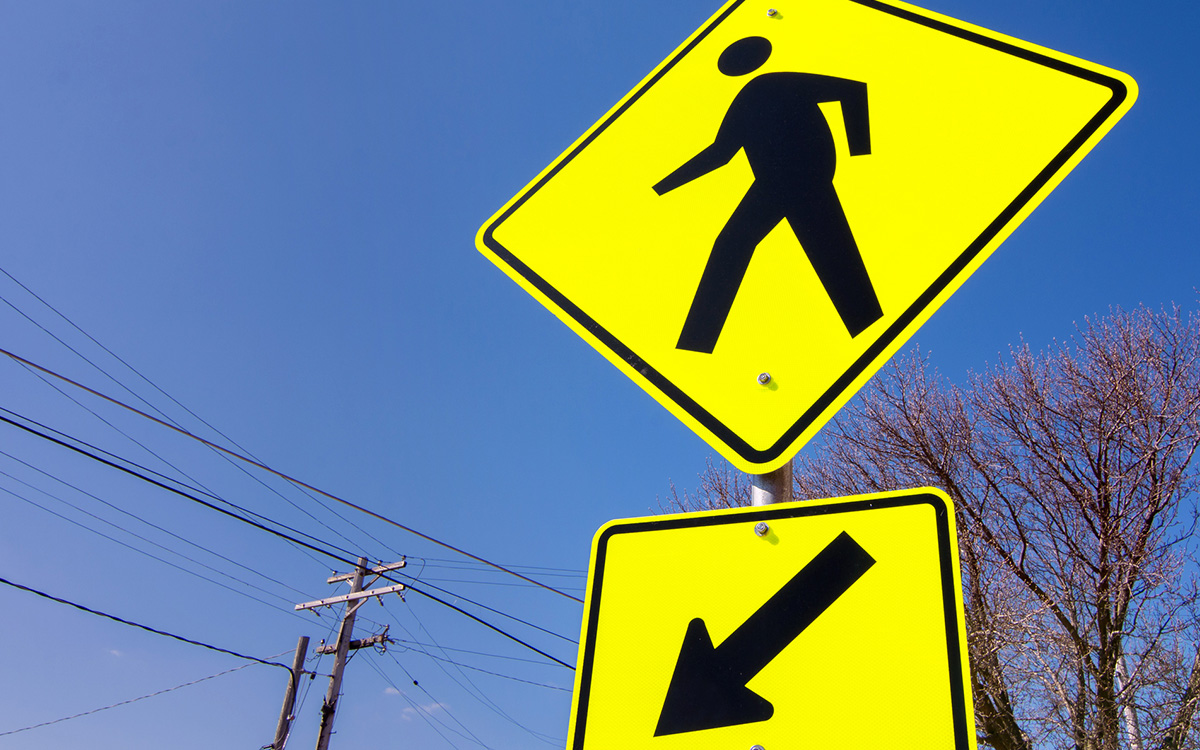 A yellow pedestrian crossing sign with a downward arrow is posted near utility lines and trees under a clear blue sky. A yellow pedestrian crossing sign with a downward arrow is posted near utility lines and trees under a clear blue sky.