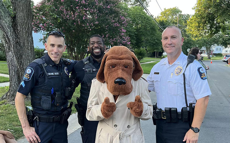 Three police officers pose with a person in a brown dog mascot costume wearing a trench coat, standing outdoors on a residential street.