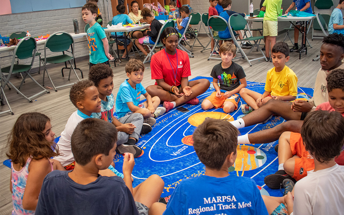 A group of children and two adults sit in a circle on a solar system rug in a classroom, engaging in an activity together. Other children are at tables in the background. A group of children and two adults sit in a circle on a solar system rug in a classroom, engaging in an activity together. Other children are at tables in the background.