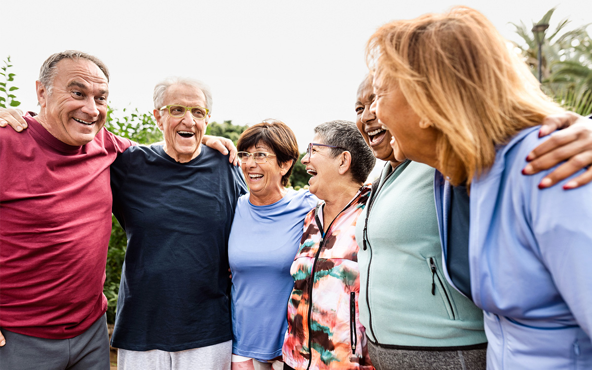 Six older adults stand outdoors in a row with arms around each other, smiling and laughing together. Six older adults stand outdoors in a row with arms around each other, smiling and laughing together.