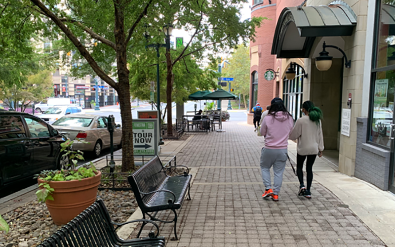 Two people walk down a tree-lined city sidewalk with benches, parked cars, and a sign for tours visible.