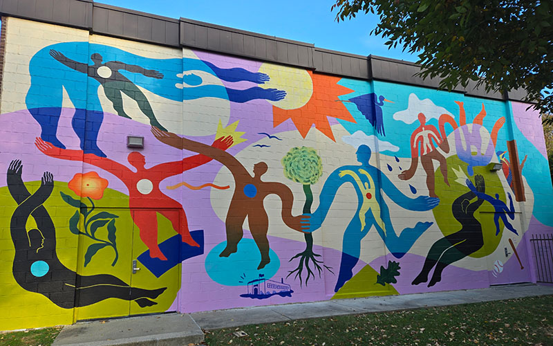 Colorful mural depicting abstract human figures, plants, and a sun on an exterior wall, with a tree partly visible in the foreground.