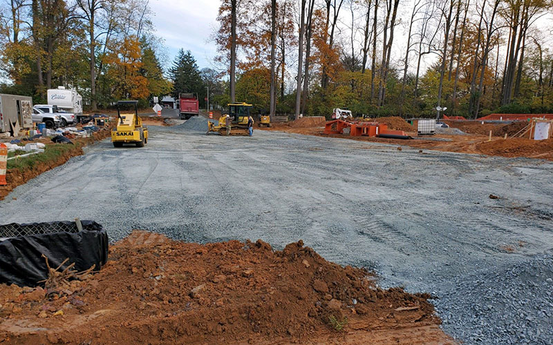 A construction site with heavy machinery leveling a gravel surface, surrounded by dirt piles, equipment, and trees in the background.