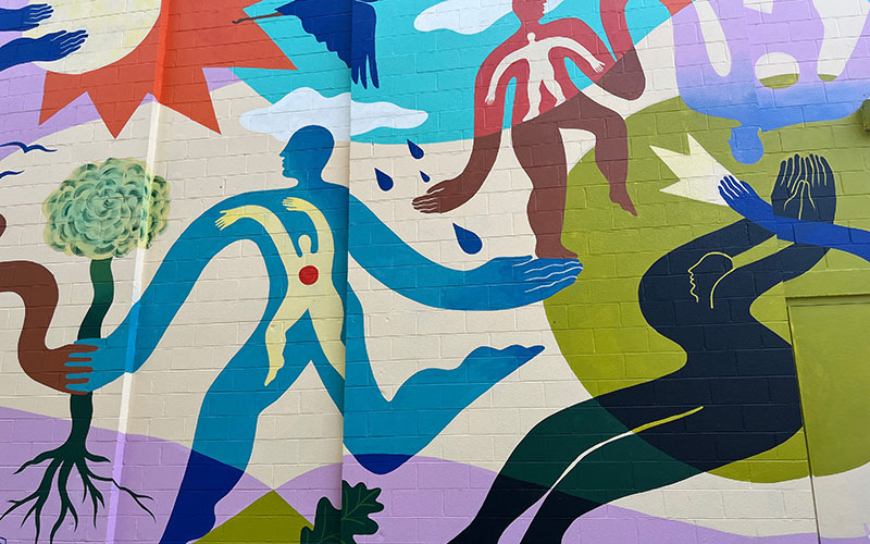 Colorful mural featuring abstract human figures, hands, and organic shapes with various symbols including a tree, droplets, and a sun, painted on a brick wall.