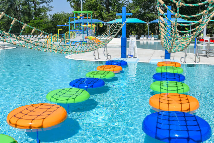 A pool obstacle course with floating orange, green, and blue stepping pads and rope bridges, set in an outdoor water park with play structures and umbrellas in the background.