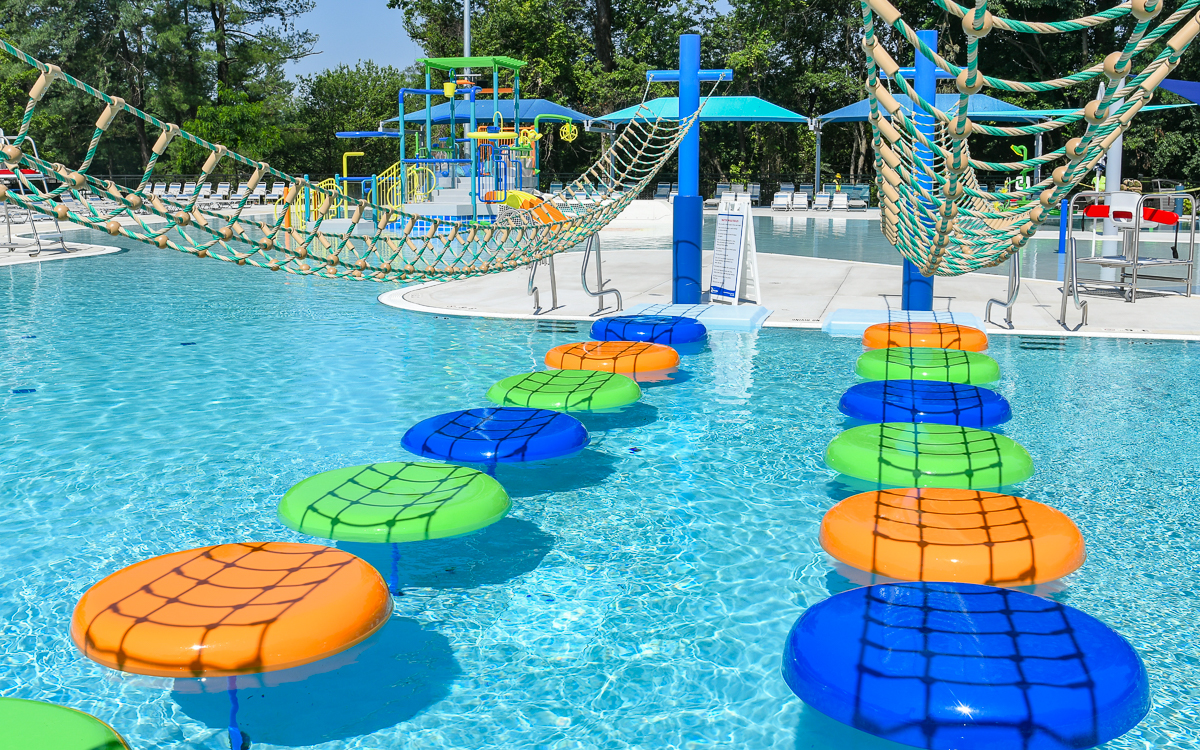 A pool obstacle course with floating orange, green, and blue stepping pads and rope bridges, set in an outdoor water park with play structures and umbrellas in the background.
