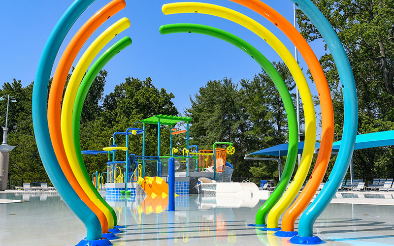 Colorful water play structures and splash arches are set in a shallow outdoor pool area with trees and blue sky in the background.