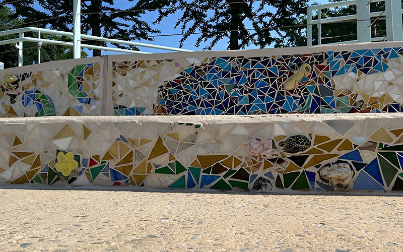 Colorful mosaic tiles decorate the side of outdoor concrete steps, featuring geometric patterns and images of sea creatures. Trees are visible in the background.