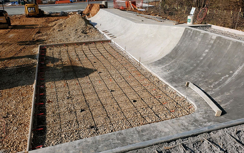 Concrete skatepark under construction showing a partially completed ramp and a section with exposed rebar grid over gravel, ready for pouring concrete. Construction equipment visible in background.