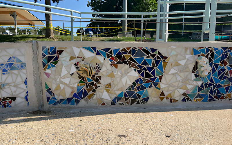 A concrete wall decorated with a mosaic of colorful, irregularly shaped tiles in blue, white, gold, and brown. Grass, railing, and caution tape are visible in the background.