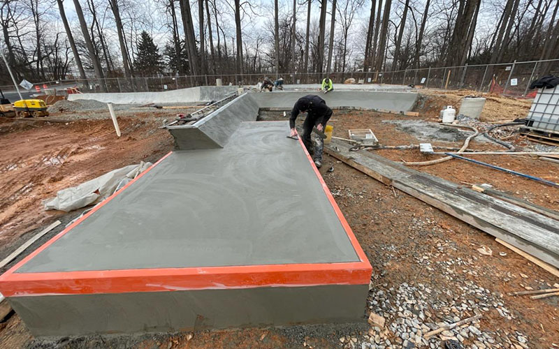 A worker smooths freshly poured concrete on a rectangular structure at a construction site surrounded by trees and building materials.