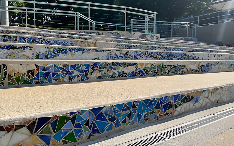 Outdoor steps decorated with a mosaic of colorful, broken tiles in blue, green, and white, next to a beige ramp and metal railings.