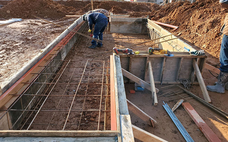 Construction workers preparing rebar and wooden forms for a concrete foundation at a building site with tools and materials scattered nearby.