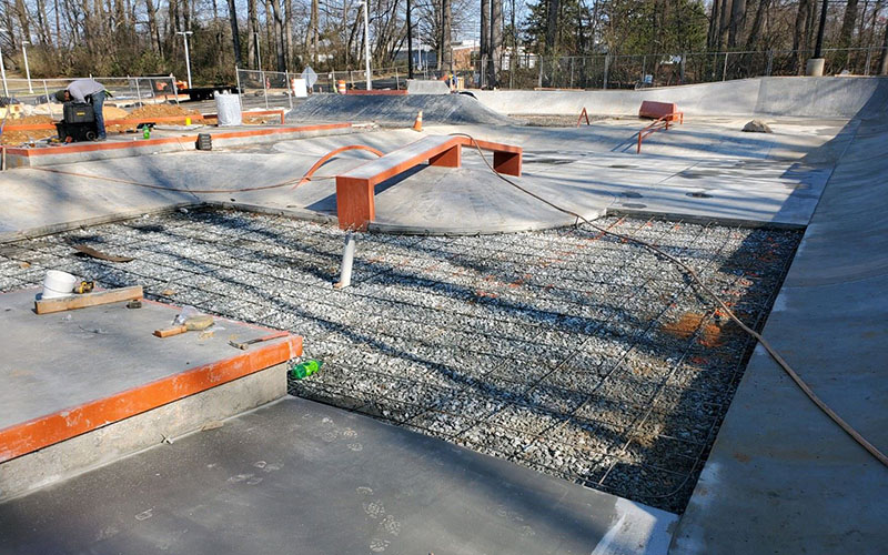 A concrete skatepark under construction with exposed rebar, gravel, and partially built ramps and features.