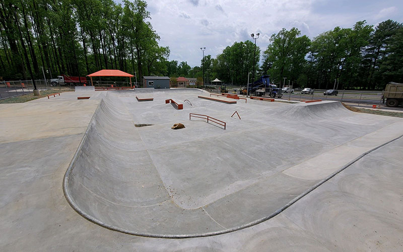 A concrete skatepark with ramps, rails, and ledges, surrounded by trees and some construction equipment in the background.