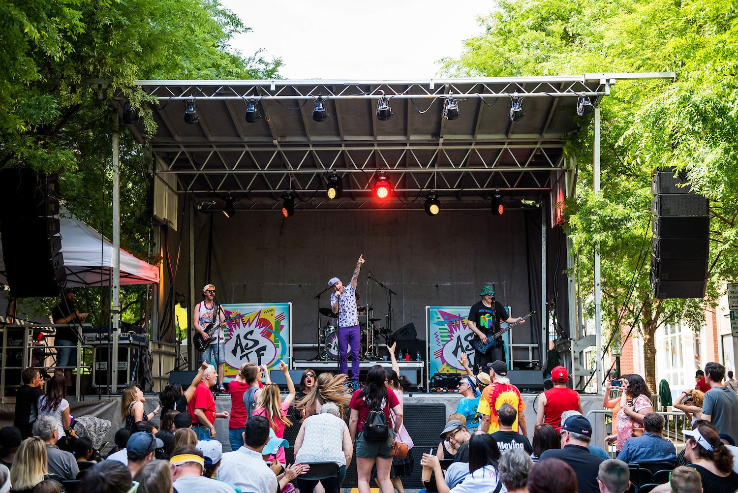 A band performs on an outdoor stage while a crowd watches and dances; trees and buildings frame the background.