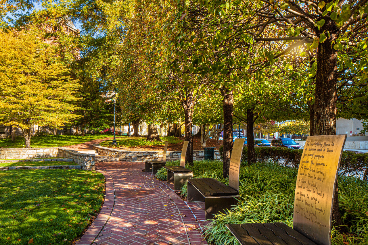 A brick pathway curves through a landscaped park with trees, benches, and inscribed plaques in sunlight.