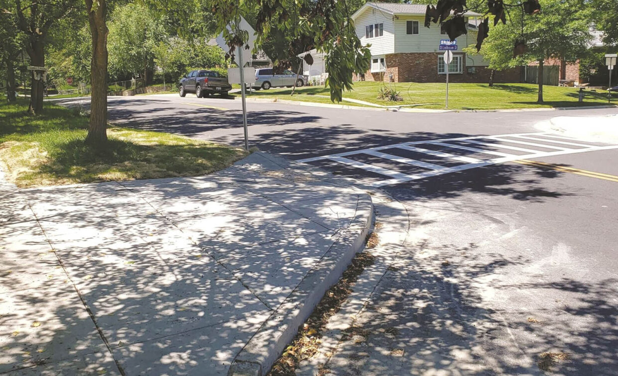 A residential street with a crosswalk, sidewalk, trees casting shadows, parked cars, and houses in the background on a sunny day.