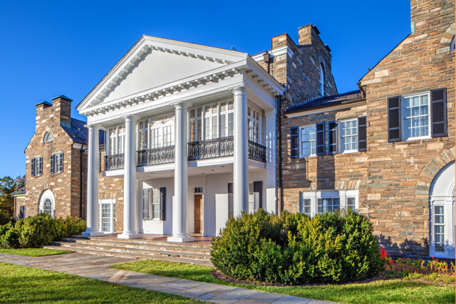 Large stone mansion with white columns, a portico, black shutters, and manicured bushes in front, photographed under clear blue sky.