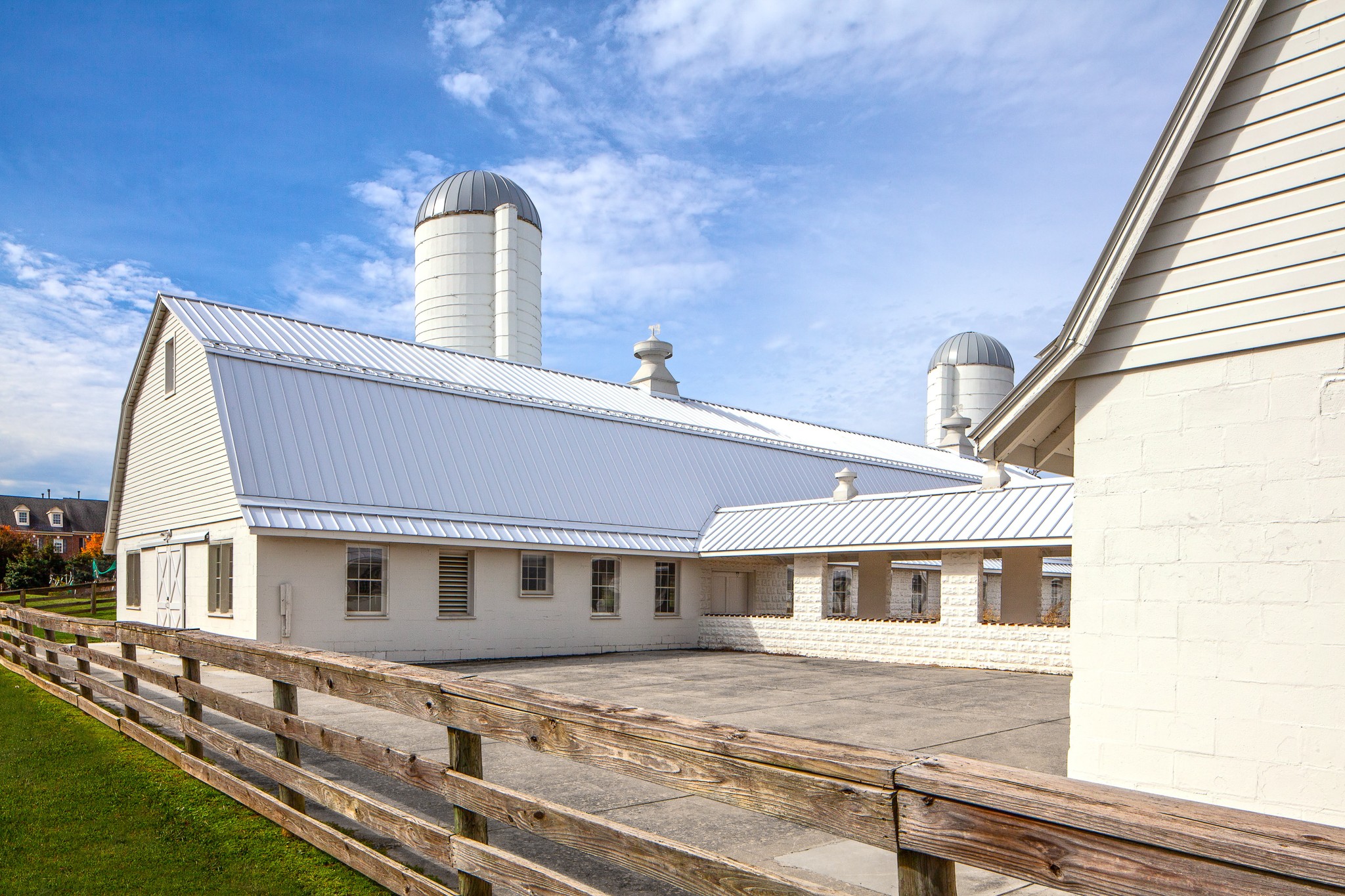 White barn buildings with metal roofs and silos, surrounded by a wooden fence under a partly cloudy sky.
