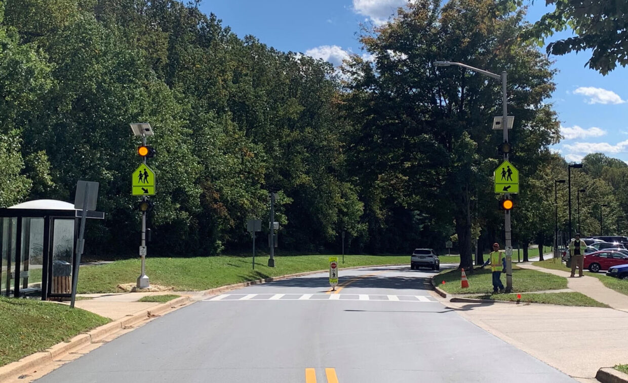 A crosswalk with flashing pedestrian crossing signs, a bus stop shelter on the left, and two people standing near the sidewalk on a sunny day.