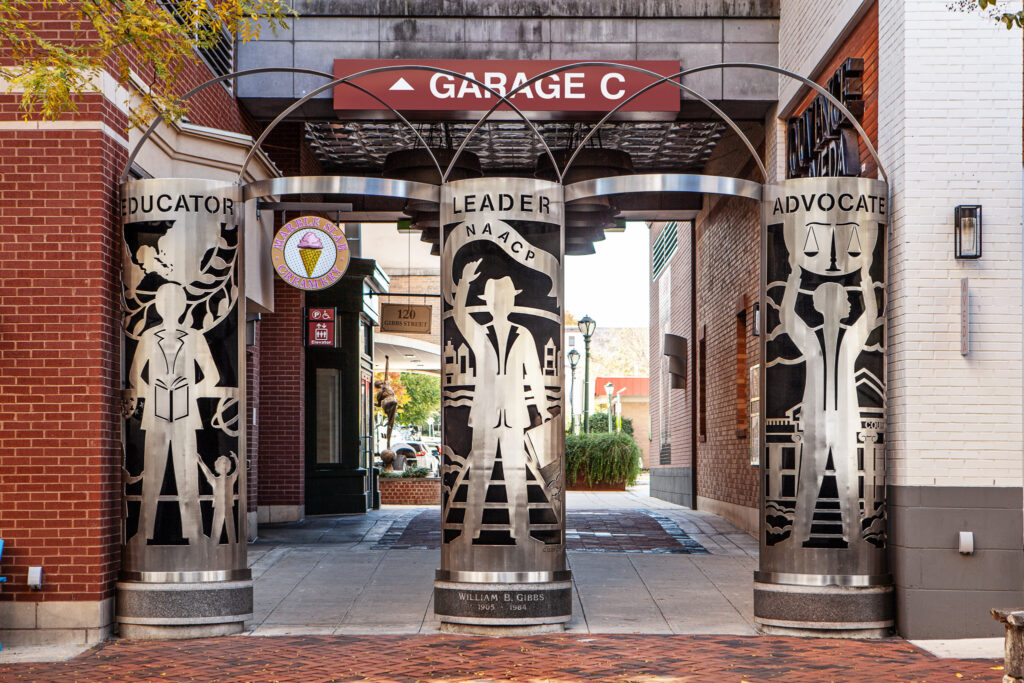 Three metal sculptures labeled "Educator," "Leader NAACP," and "Advocate" stand under an arch in a brick-paved walkway near a sign for "Garage C.