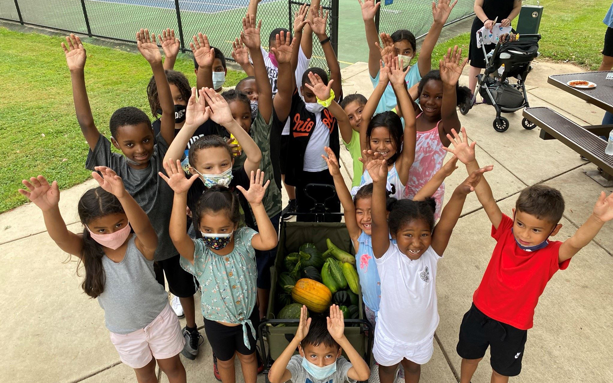A group of children outdoors stand around a container of vegetables, raising their hands and smiling at the camera.