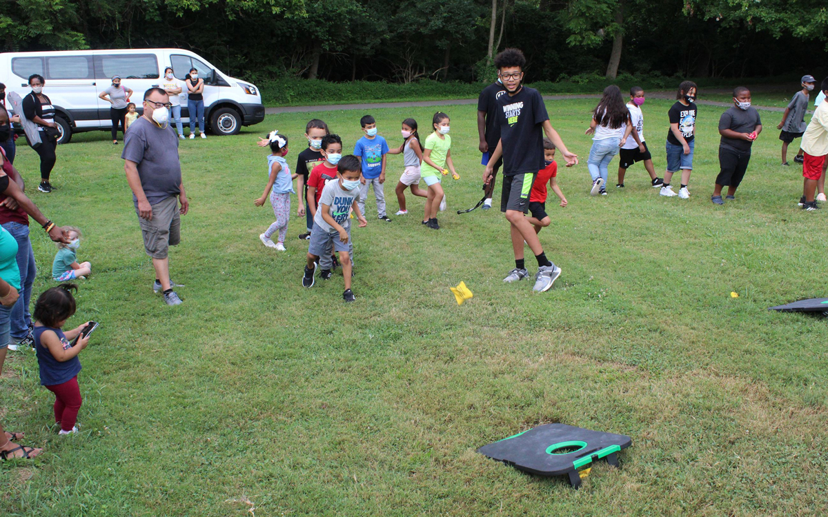A group of children and adults play an outdoor lawn game on grass, with some wearing masks; a white van and trees are in the background.