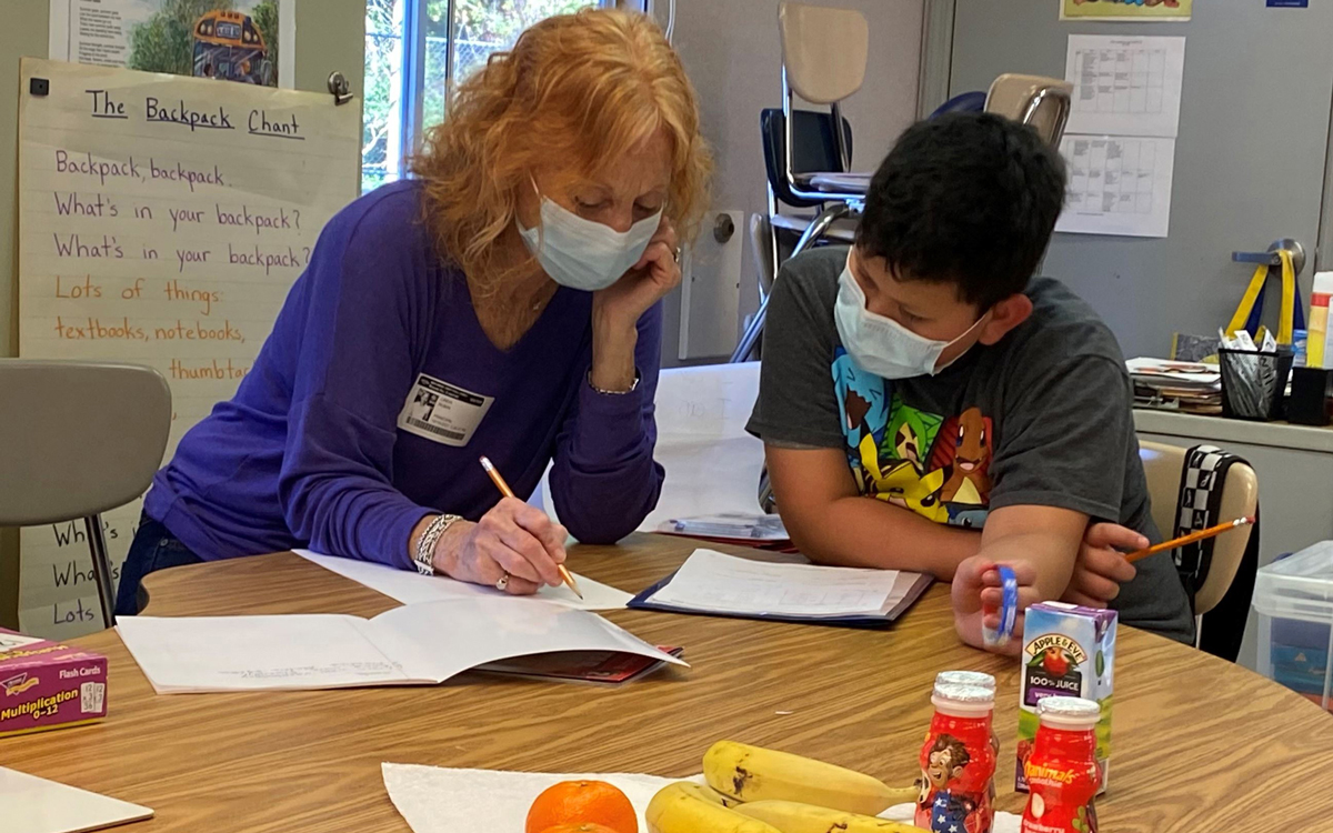 A teacher and a student, both wearing masks, sit at a table working on a worksheet together in a classroom with snacks and school supplies on the table.