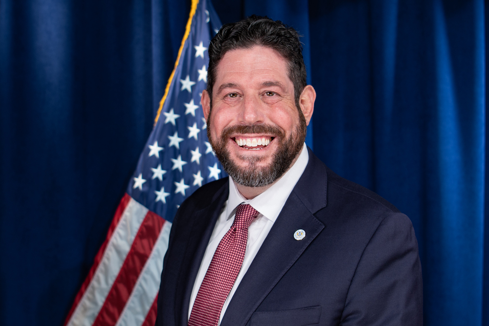 Man in a suit and red tie smiling, standing in front of an American flag and blue curtain background.