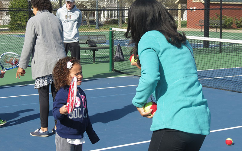 A woman holding tennis balls instructs a young girl with a racket on an outdoor tennis court; others are present in the background.