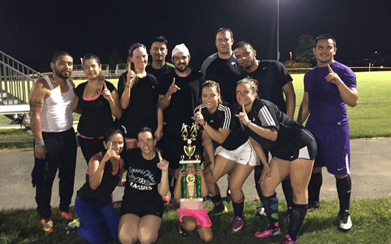 A group of eleven adults and one child pose outdoors at night, holding up one finger and displaying a large trophy on a grassy field.
