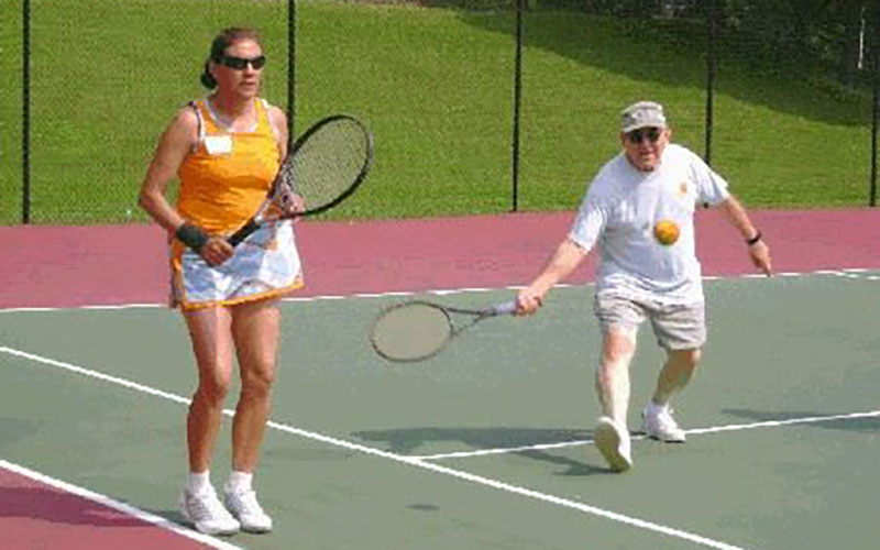 Two people playing doubles tennis on an outdoor court; one is preparing for a shot while the other is reaching to hit the ball.