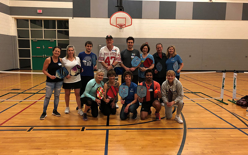 A group of people pose in a gymnasium, some kneeling and some standing, holding pickleball paddles in front of an indoor court and basketball hoop.