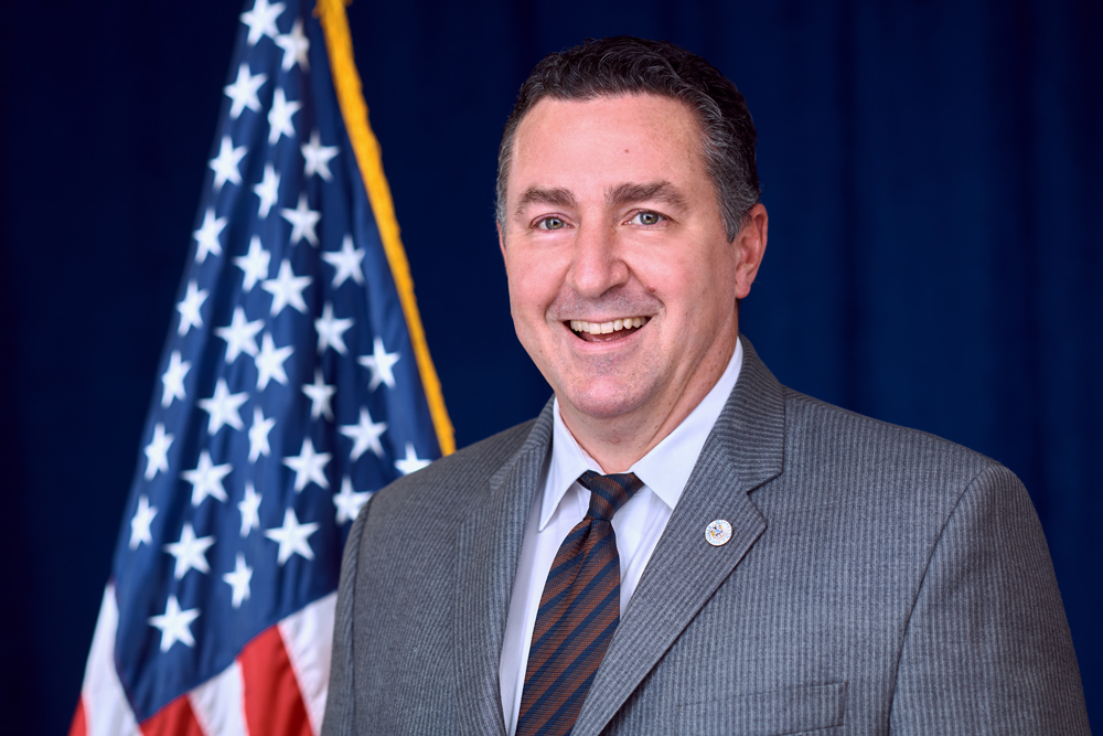 A man in a gray suit and striped tie smiles while standing in front of an American flag and a dark blue backdrop.