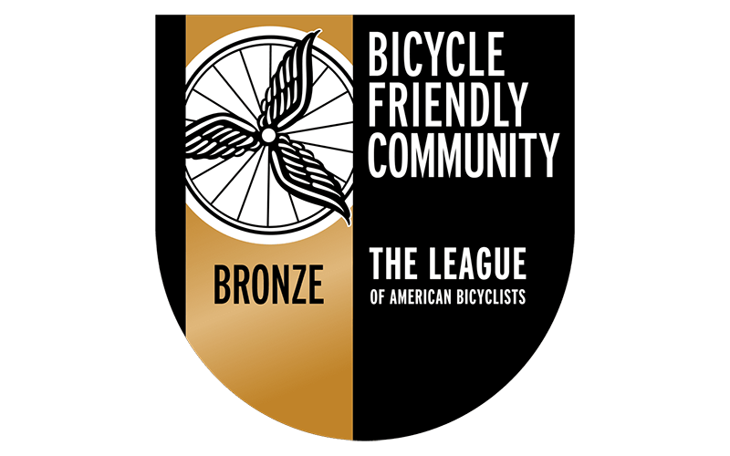 A badge with a bicycle wheel graphic, labeled "Bicycle Friendly Community Bronze" and "The League of American Bicyclists.